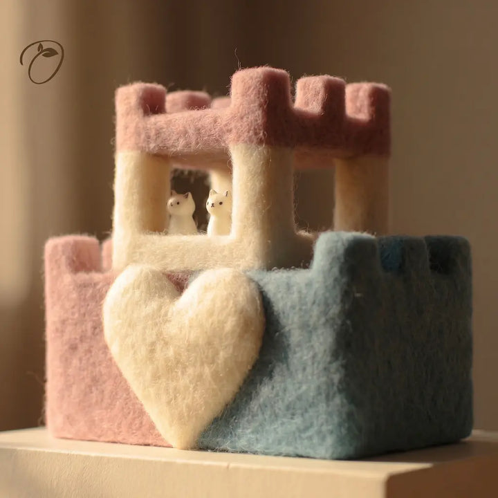 A pastel felt decorative structure resembling a small house or shelf, with a large heart-shaped cushion in front. The top section is cream with a pink roof, and the lower left section is pink while the lower right is muted blue-green.