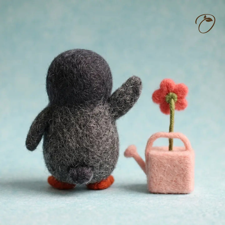 A small felt penguin figurine seen from the back, with orange feet, standing beside a pink felt watering can holding a red flower, on a soft light blue background.