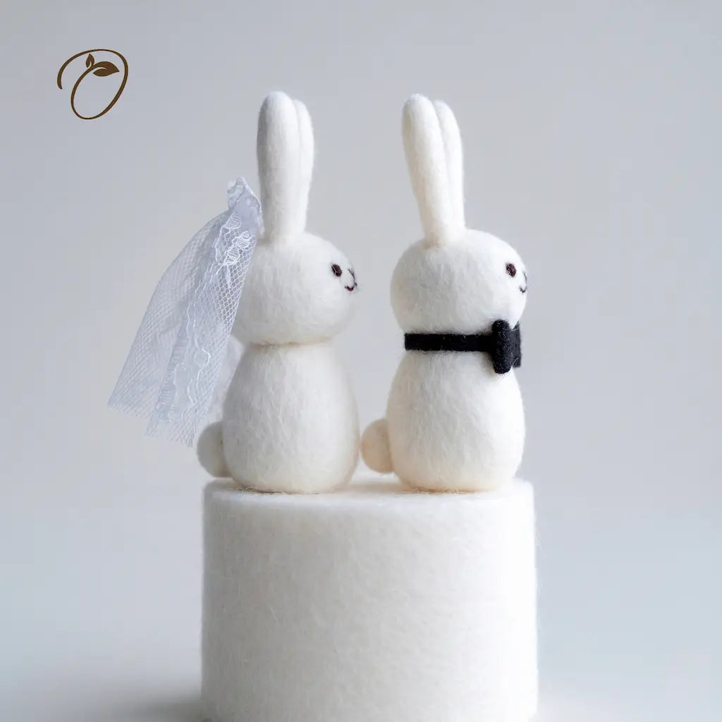 Two white rabbit figurines, one with a veil and bow tie, on a white stand against a light gray background.