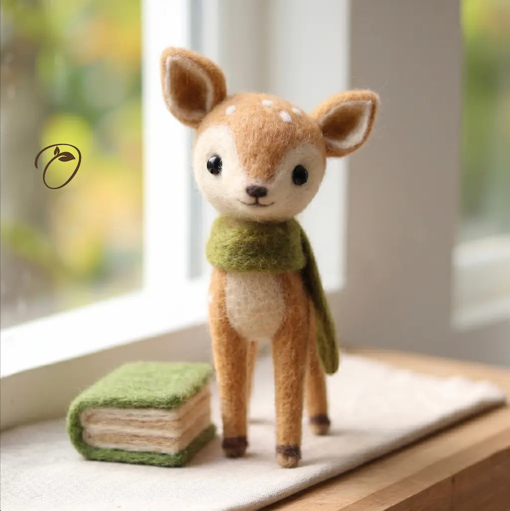 A cute needle-felted toy deer standing on a window sill next to a small felted book, wearing a green scarf.