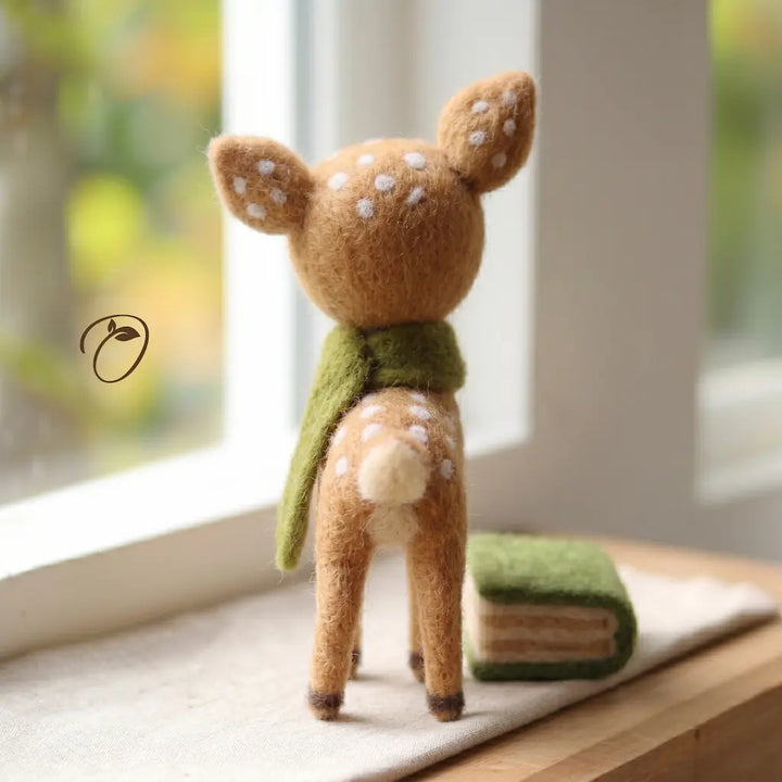 A small felt deer figurine stands on a windowsill, facing away from the camera. The deer is tan with white spots and wears an olive green scarf.