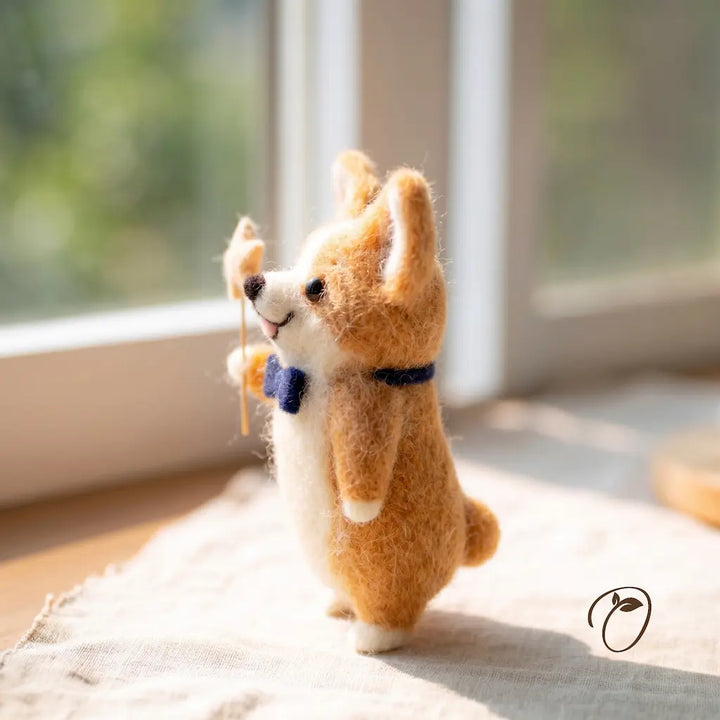 A handcrafted felt needle art corgi figurine standing on its hind legs, holding a small twig branch, set on a light fabric surface in front of a softly blurred window.