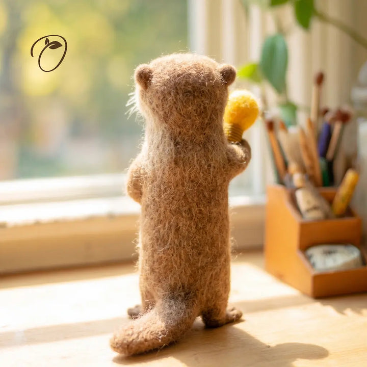 A small felted animal figure, resembling a beaver or otter, stands on its hind legs on a wooden surface near a window. The figure is brown with a soft, fuzzy texture and holds a yellow brush in one paw.
