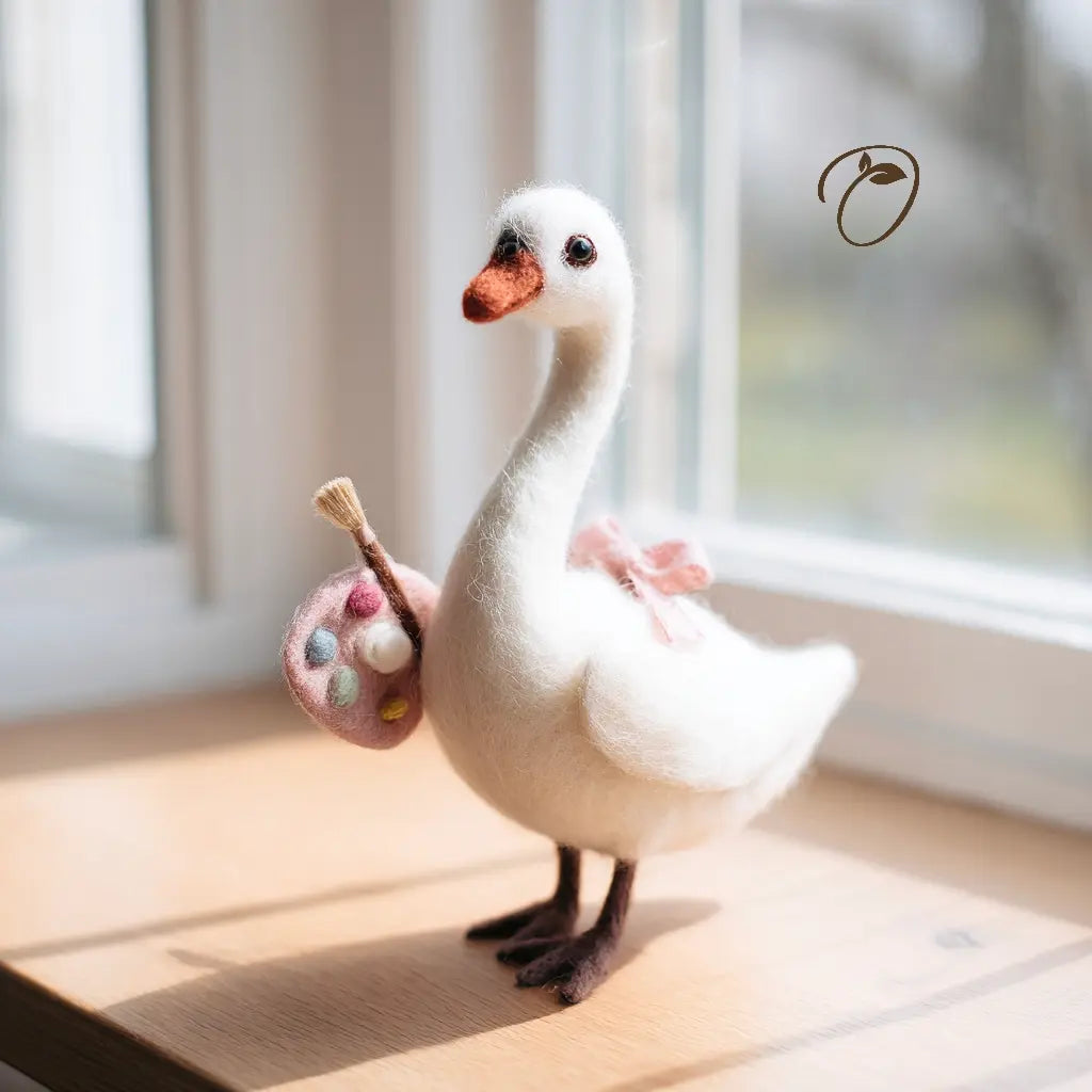 A white felted wool swan figurine standing on a wooden windowsill, holding a pink painter's palette with a paintbrush. The figurine has a pink bow on its back and dark brown beak and legs.