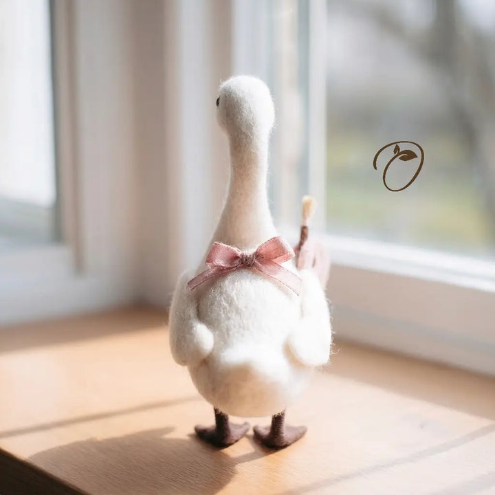 A white felt decorative goose figurine with a pink satin bow and dark brown beak and feet, standing on a wooden windowsill in soft natural light.