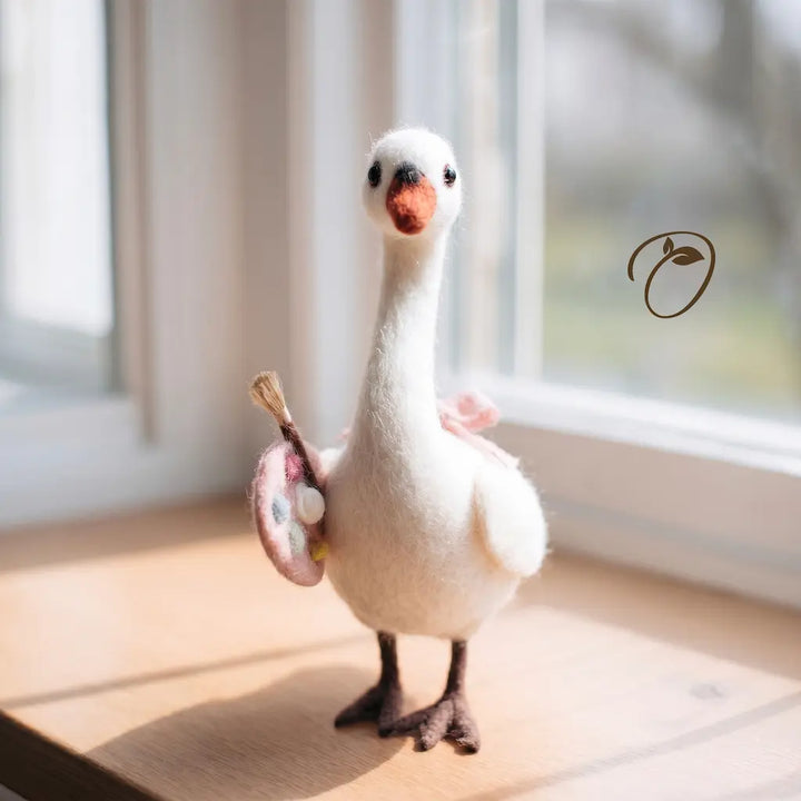 A white goose figurine standing on a wooden windowsill, holding a pink and gold paint palette. The figurine has a soft, felt-like texture, dark brown legs and feet, and a small orange beak.