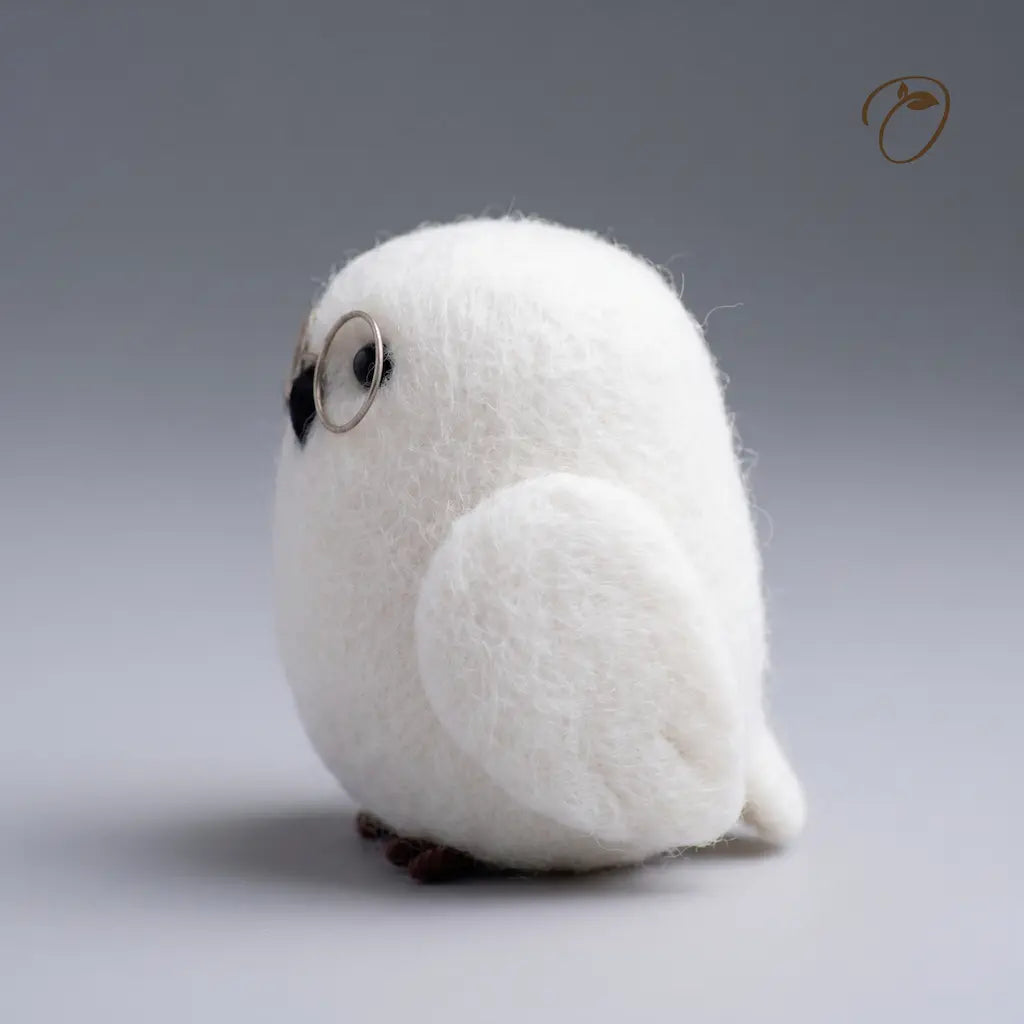 A white felt owl figurine with round wire glasses, a black beak, and small brown feet, photographed against a smooth gray gradient background.