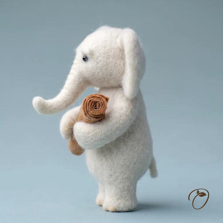 A white felted wool elephant figurine holding a rolled brown fabric flower, standing against a soft light blue-gray studio background.