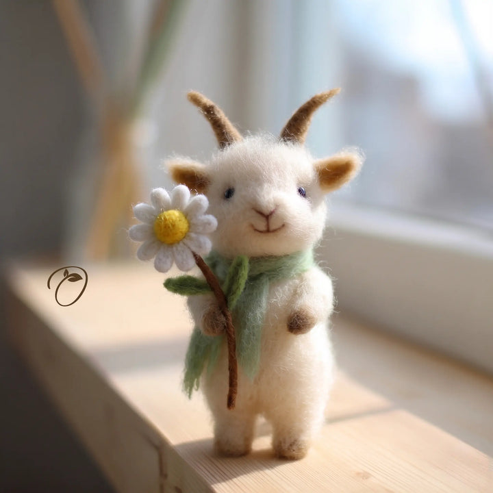 A small felted wool goat figurine with brown horns, wearing a mint green scarf and holding a white daisy with a yellow center, standing on a light wooden surface near a softly blurred window.