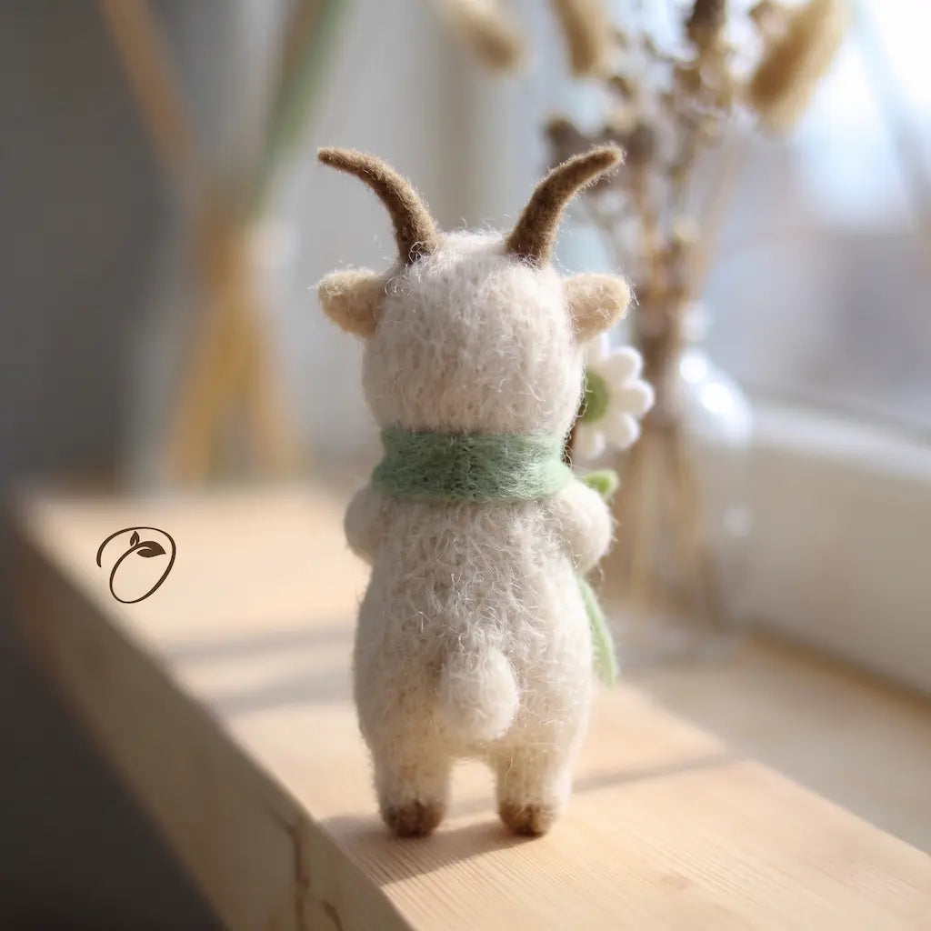 A small felted wool goat figurine stands on its hind legs on a light wooden surface, viewed from behind. The white goat has brown horns, a mint green scarf, and holds a white flower.