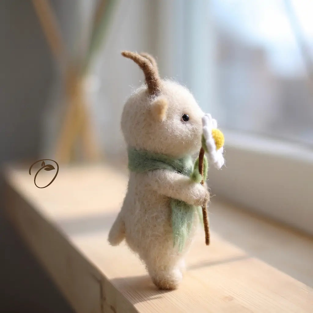 A small felted wool animal figurine with brown horns, a mint green scarf, and a white flower with a yellow center, standing on a wooden windowsill in soft natural light.