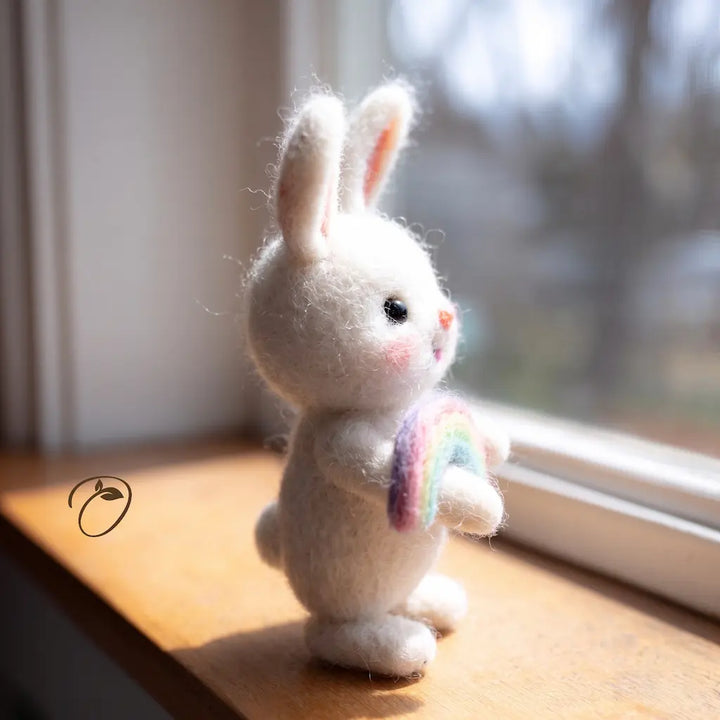 A small white felted rabbit figurine with long ears stands on a wooden windowsill, holding a pastel rainbow. The rabbit has black bead eyes and a tiny pink stitched nose.
