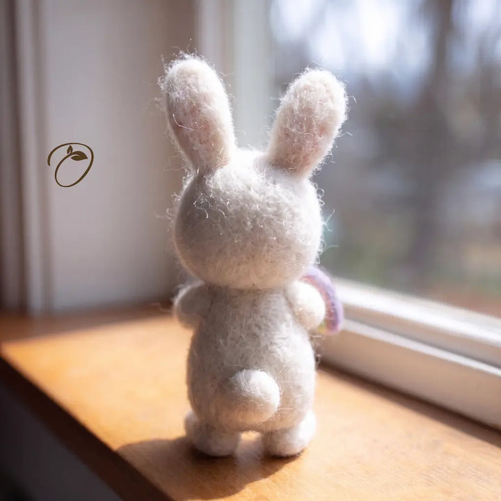 A small white felted wool bunny figurine with upright ears stands on a wooden windowsill, facing a bright window. The bunny has a soft fuzzy texture and a tiny lavender ribbon detail on one ear, with a blurred outdoor scene visible behind it.