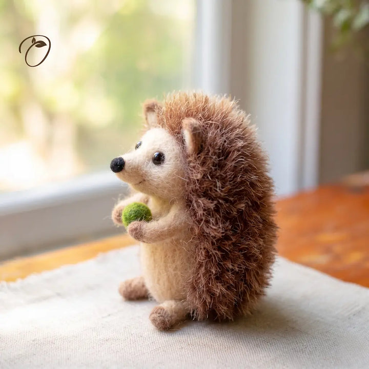 A small needle-felted hedgehog toy sits on a light fabric surface near a window, holding a green acorn. The hedgehog has brown and cream fur with a soft, fuzzy texture.