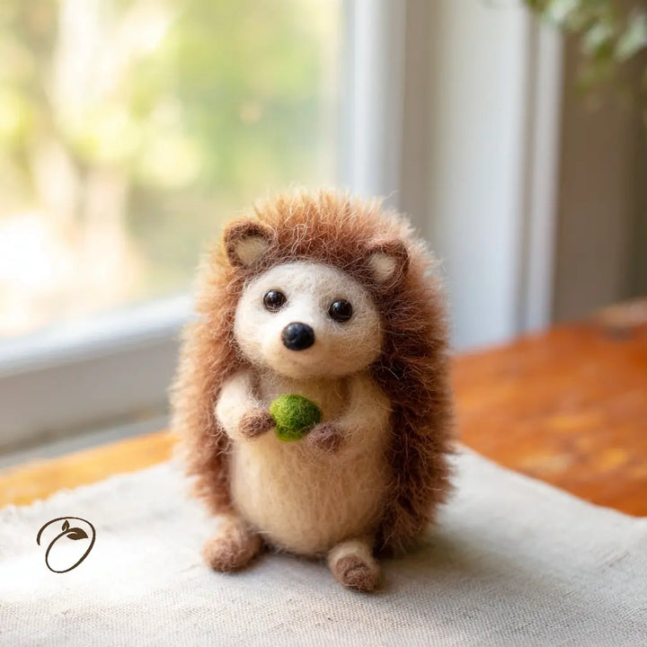 A small needle-felted hedgehog figurine with brown and cream fur, holding a green acorn, sitting on a light fabric surface near a softly blurred window.