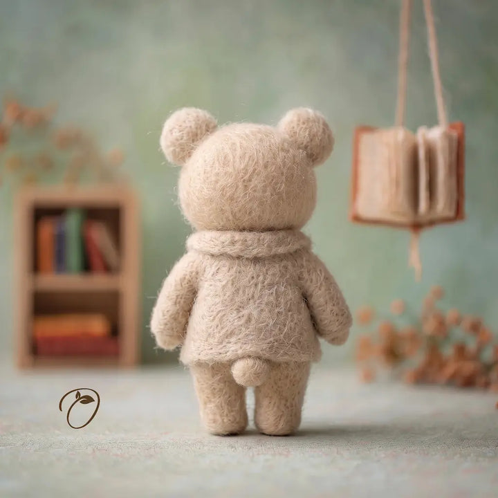 A small beige felt teddy bear viewed from behind, standing on a soft surface in a styled miniature room with blurred books and dried flowers in the background.