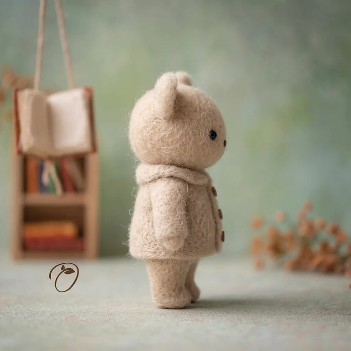 Small felted wool teddy bear figure wearing a cream-colored coat and scarf, shown from the back in a softly styled miniature scene with a tiny bookshelf and dried flowers in the background.