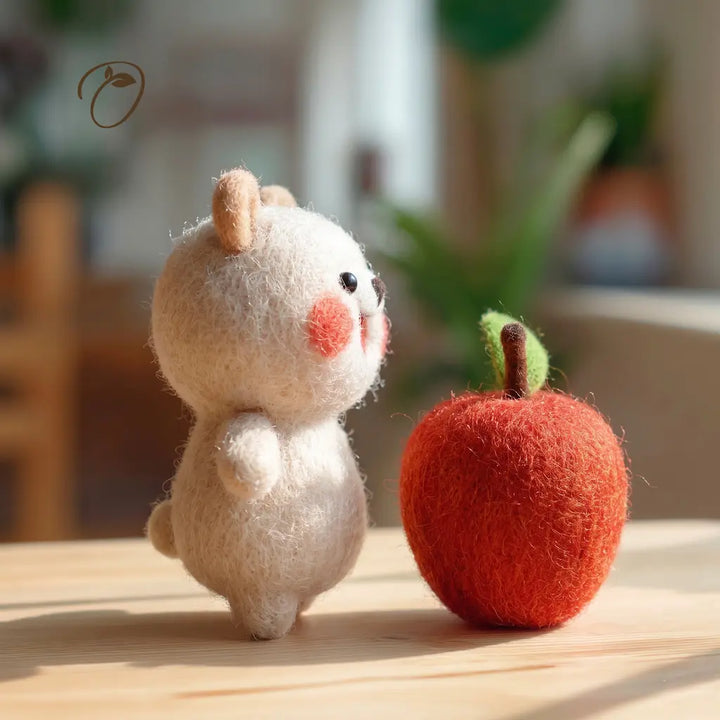 A small felted animal figurine in cream with pink cheeks stands beside a red felted apple on a light wooden surface, with a softly blurred indoor background.
