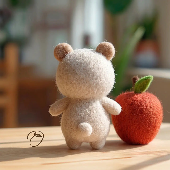 A small felt teddy bear figurine shown from the back, standing on a wooden surface next to a red apple prop. The background is a softly blurred indoor setting.