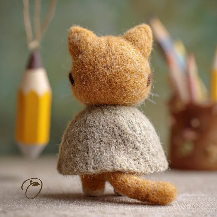 Small brown cat figurine wearing a beige dress with pencils in the background.