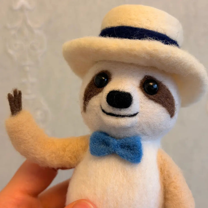 Sloth figurine wearing a hat and bow tie held by a hand against a plain background