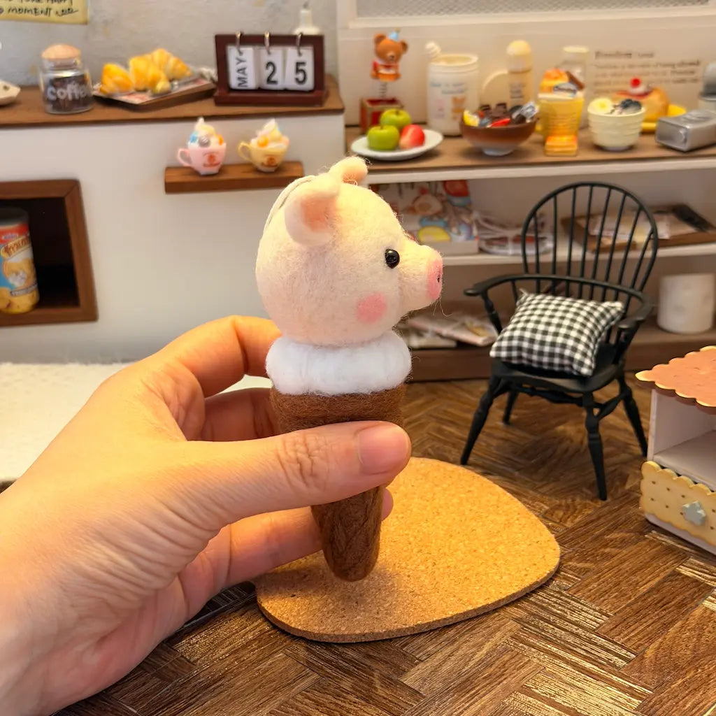 Hand holding a small pig figurine on an ice-cream cone with a miniature room in the background.