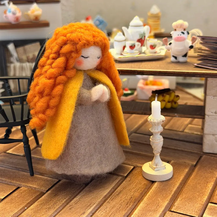 Felted doll with orange hair and yellow scarf on a wooden floor with a candle and tea set in the background.