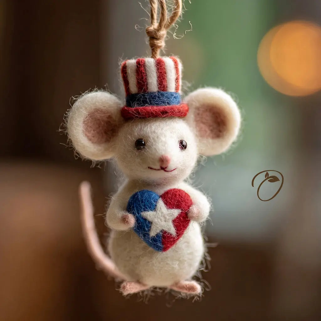 A felted wool mouse ornament wearing a red, white, and blue Uncle Sam hat and holding a red, white, and blue heart, hanging against a softly blurred warm brown background.