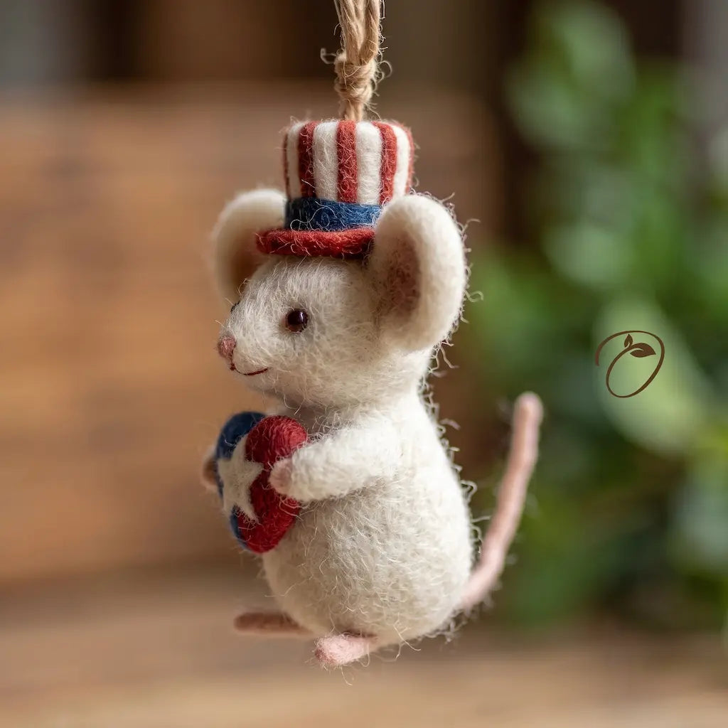 A felted wool mouse ornament dressed as Uncle Sam, wearing a red, white, and blue striped top hat and holding a small red, white, and blue star-patterned heart, hanging against a softly blurred indoor background.