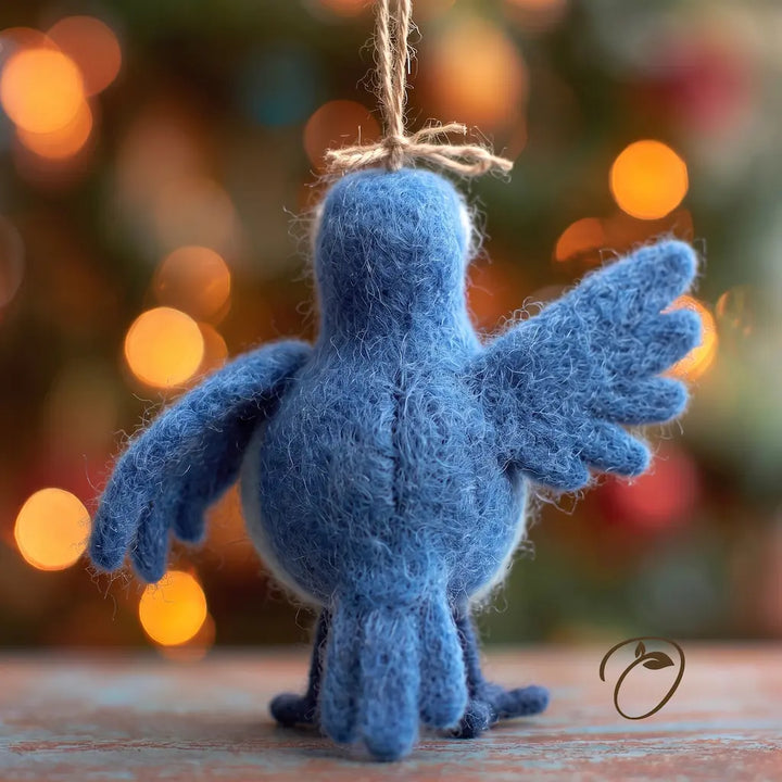 A blue felt bird ornament with wings spread, hanging by a twine loop, photographed on a wooden surface with a blurred festive background of warm lights.