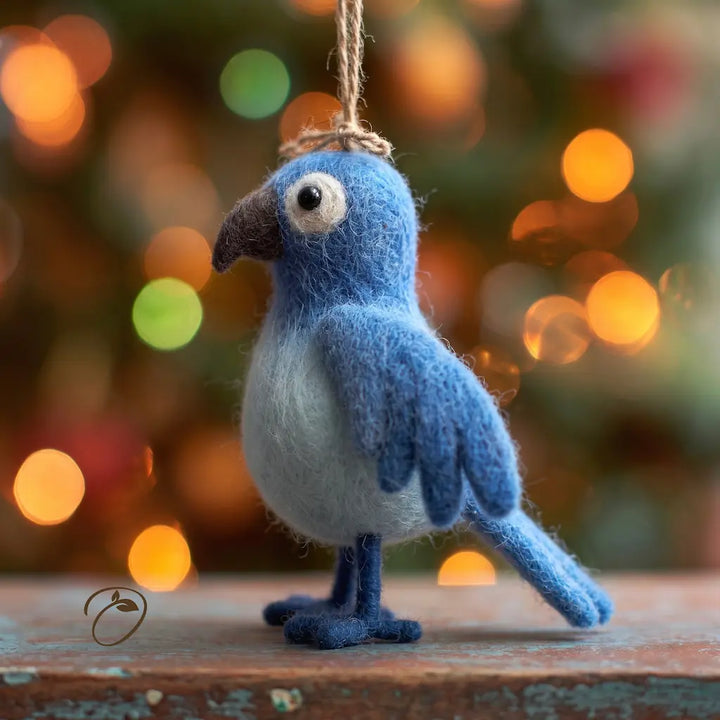 A blue and white felted wool bird ornament hanging from a twine loop, standing on a rustic wooden surface with a blurred Christmas tree and warm bokeh lights in the background.