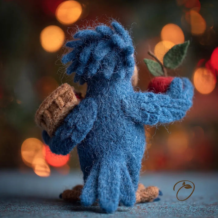 A blue fuzzy felt bird figurine holding a brown acorn in one hand and a red apple with green leaves in the other, standing on a blue surface with a warm blurred bokeh background.