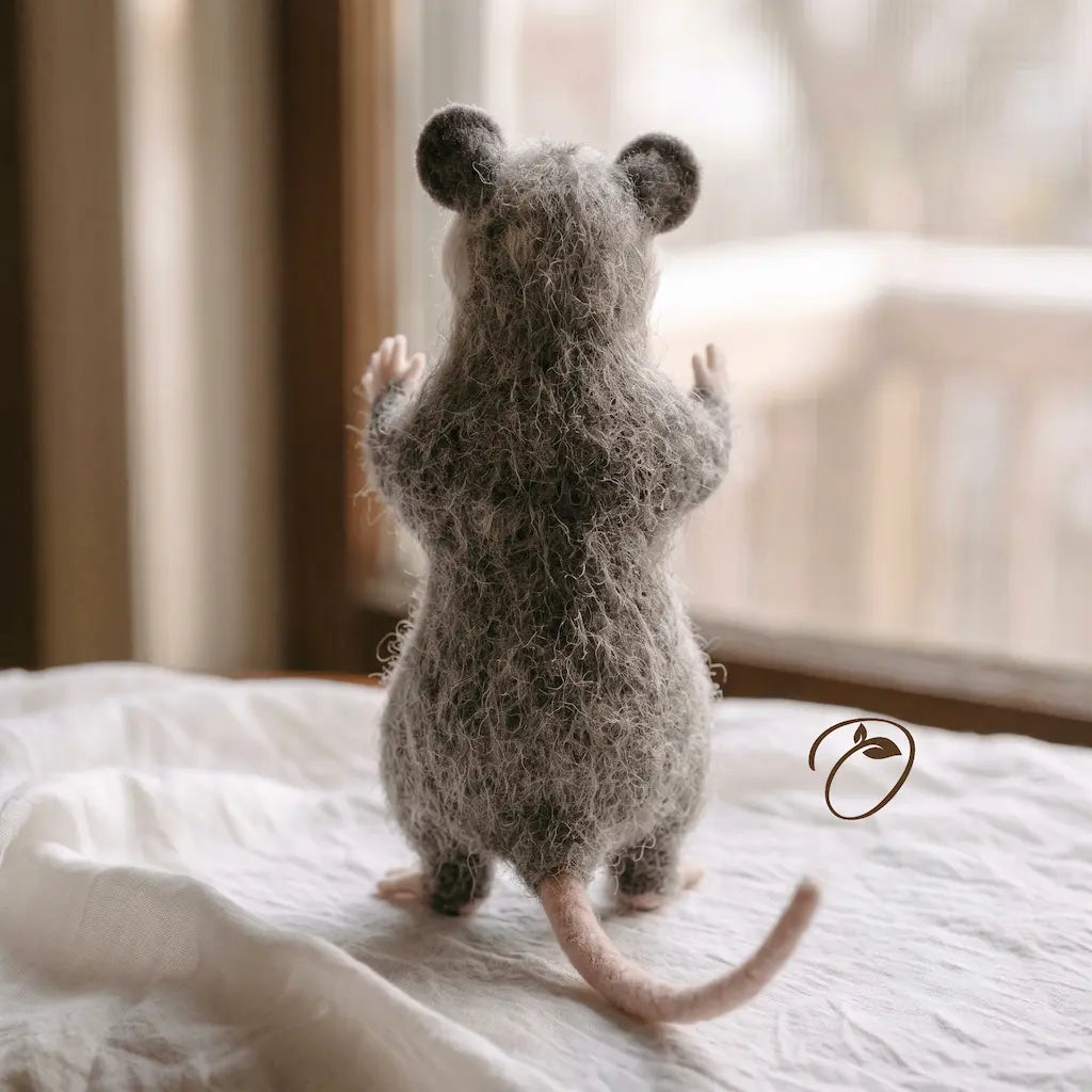 A fluffy gray felted wool mouse figurine stands on its hind legs on a white bed, facing a softly blurred window.
