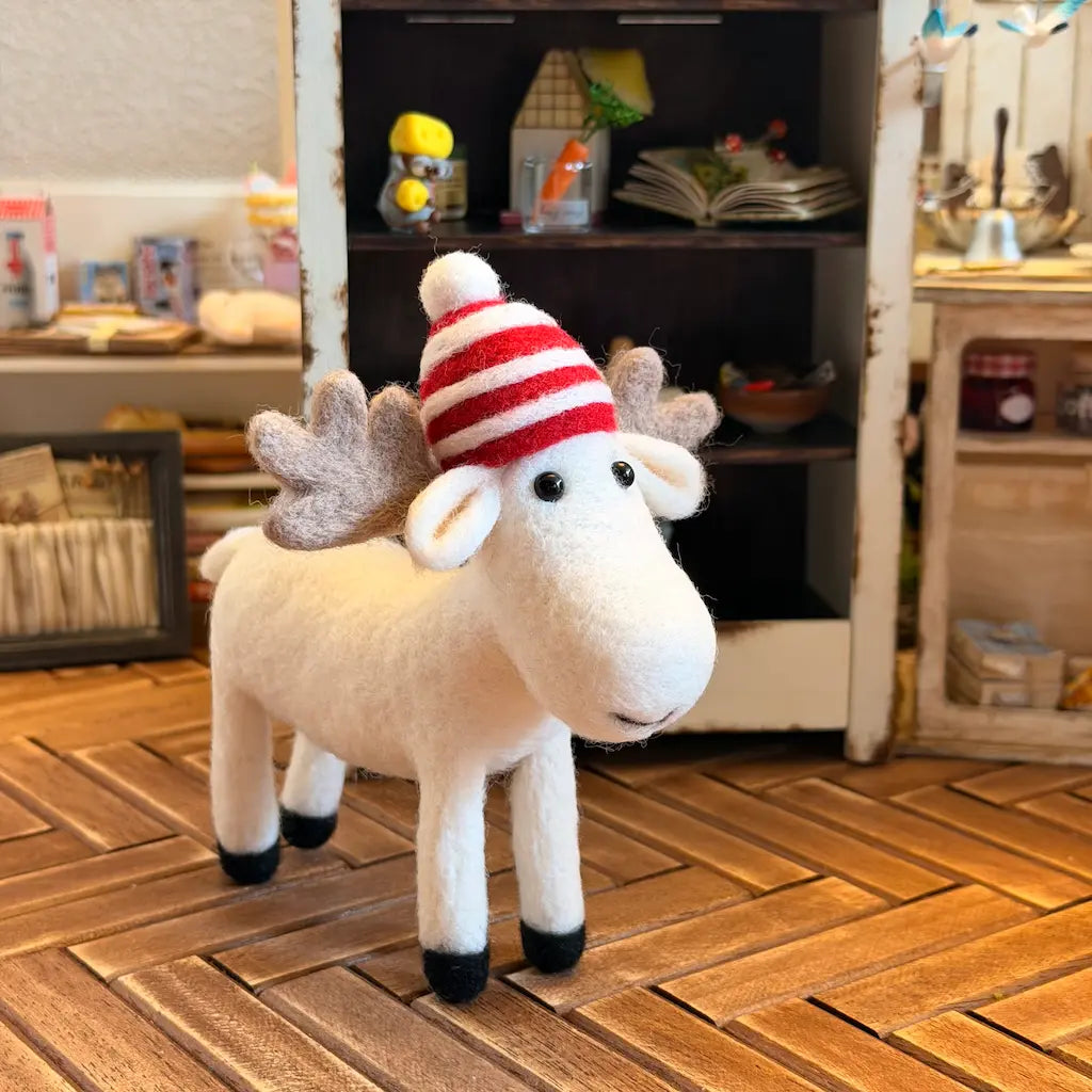 A small white moose figurine with gray antlers and a red-and-white striped knit hat, standing on a wooden floor in a cozy indoor room.