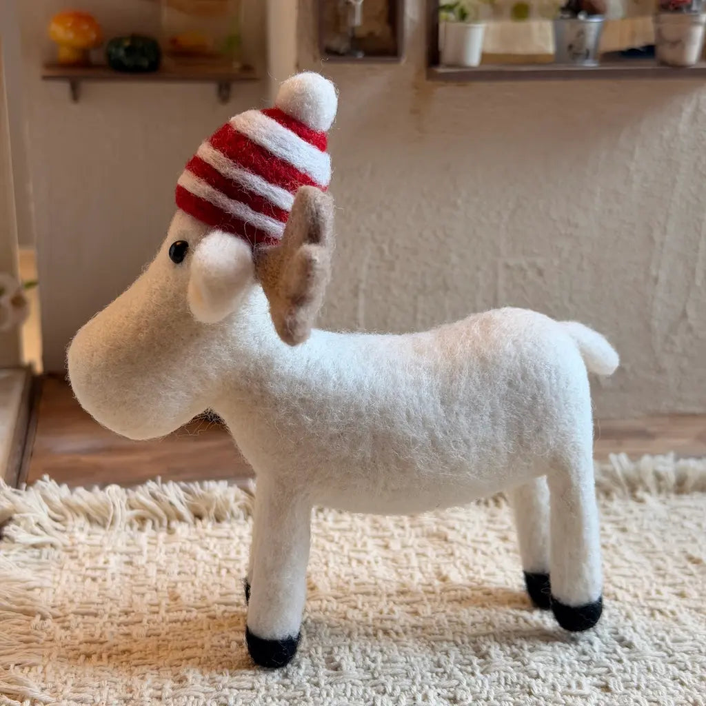 A small white moose figurine with gray antlers and a red-and-white striped knit hat, standing on a wooden floor in a cozy indoor room.