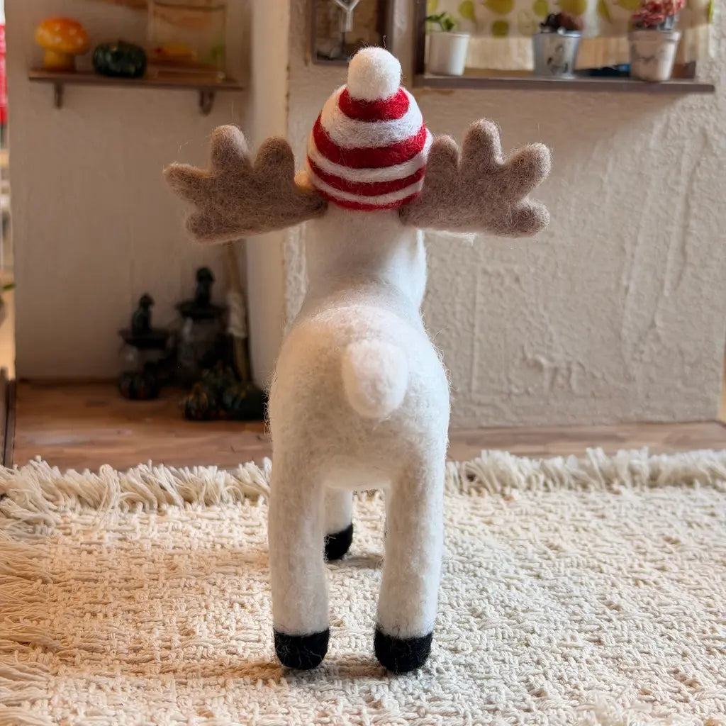 A small white moose figurine with gray antlers and a red-and-white striped knit hat, standing on a wooden floor in a cozy indoor room.
