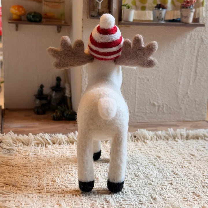 A small white moose figurine with gray antlers and a red-and-white striped knit hat, standing on a wooden floor in a cozy indoor room.