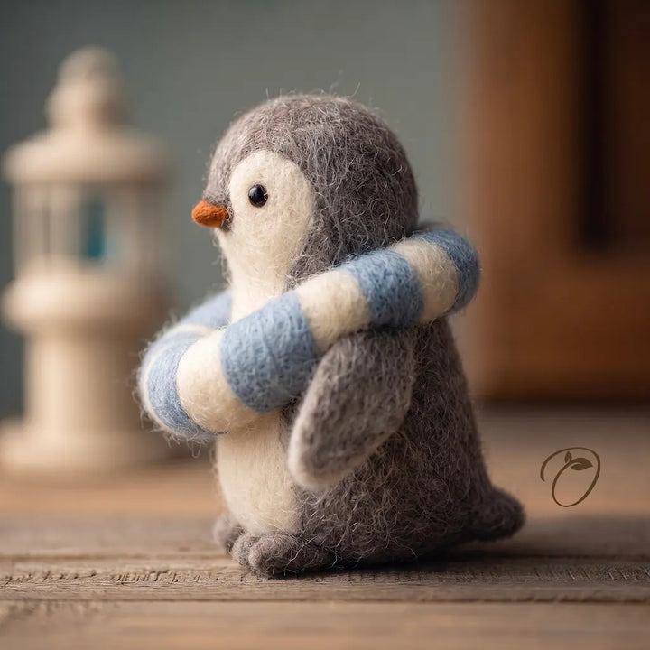 A small felted penguin figurine with gray and white feathers, wearing a blue and cream striped scarf, standing on a wooden surface with a softly blurred indoor background.