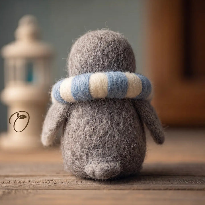 A small gray felt penguin figurine seen from the back, wearing a blue and cream striped scarf, sitting on a wooden surface with a softly blurred indoor background.