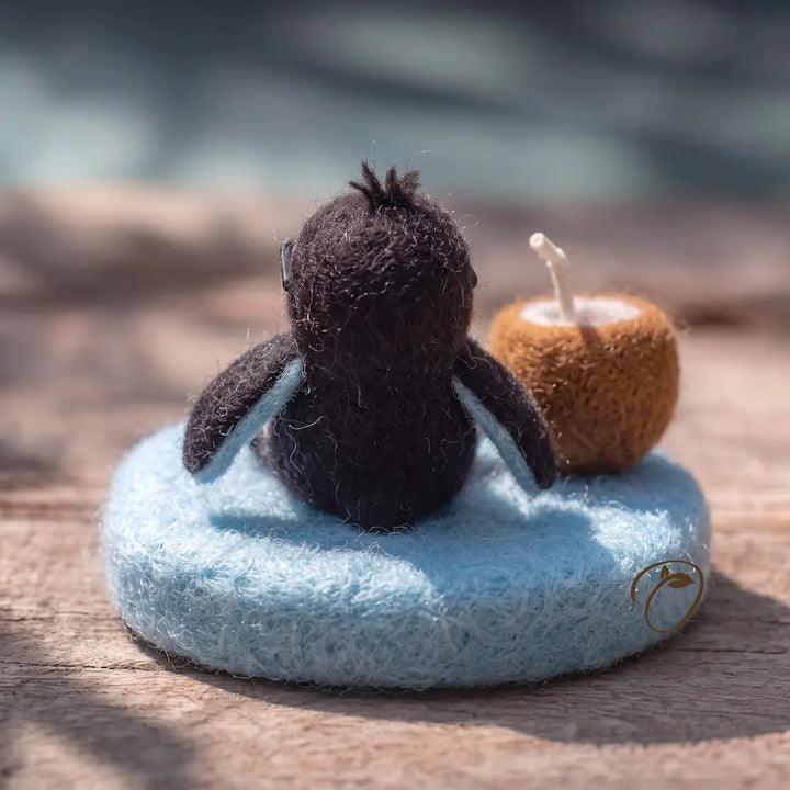A small felted wool penguin figurine sits on a round light blue base on a wooden surface, with a brown candle behind it. The photo is shot in soft natural light with a blurred outdoor background.