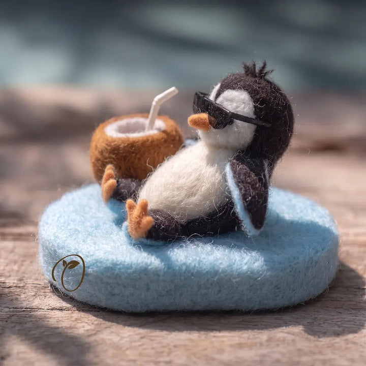 A small felted wool penguin figurine wearing black sunglasses and reclining on a round light blue cushion, with a brown coconut drink in front. The scene is photographed on a wooden surface with a softly blurred outdoor background.