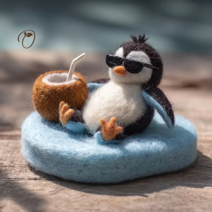 A felt penguin figurine wearing black sunglasses and sitting on a light blue round base, next to a brown coconut drink with a white straw, placed on a wooden surface with a blurred outdoor background.