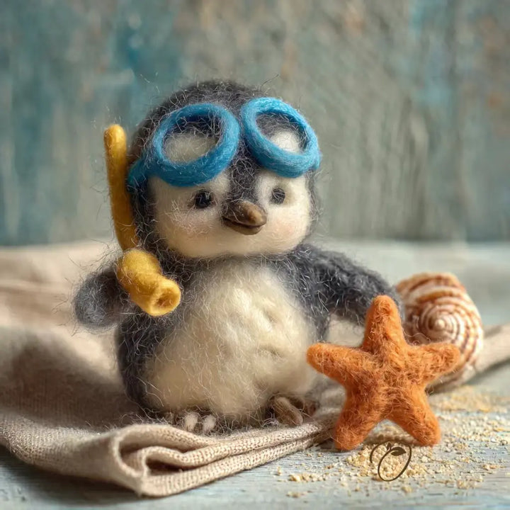 A needle-felted penguin toy wearing blue swim goggles and holding a yellow snorkel, posed on a beige fabric surface with a starfish, seashell, and sandy texture against a rustic blue background.