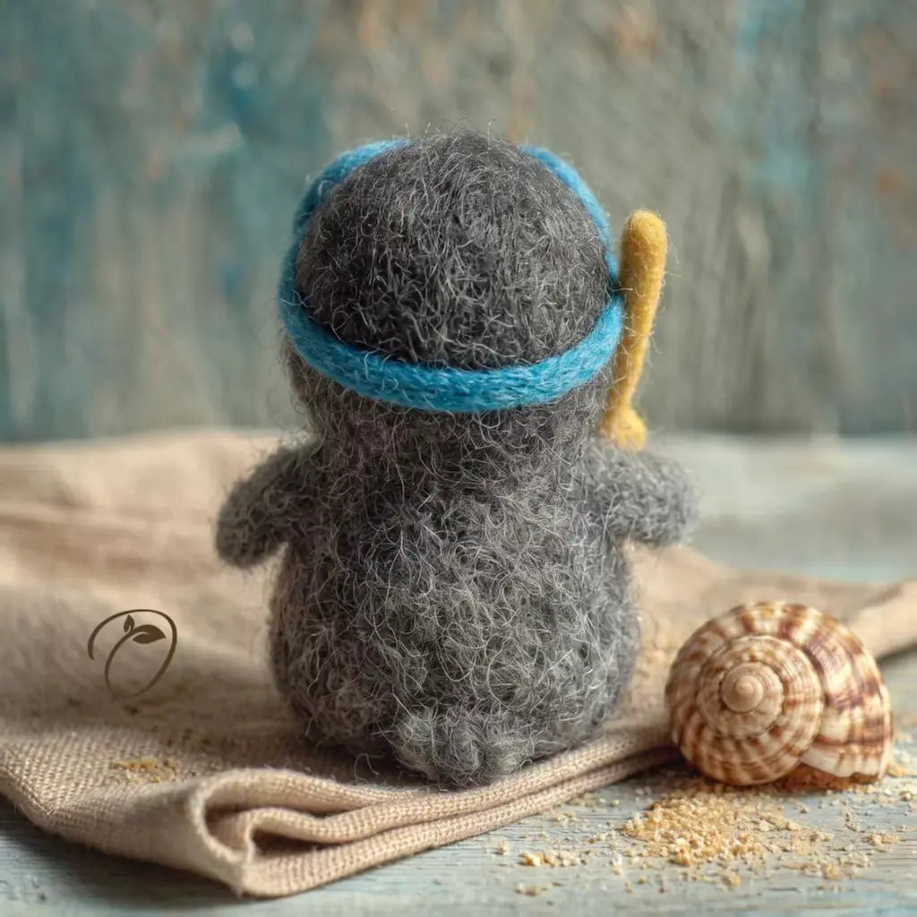 A small gray felt penguin figurine sits on a beige fabric surface, facing away from the camera. It wears a bright blue headband and holds a yellow paddle, with a striped seashell and sand-like crumbs nearby against a textured blue background.