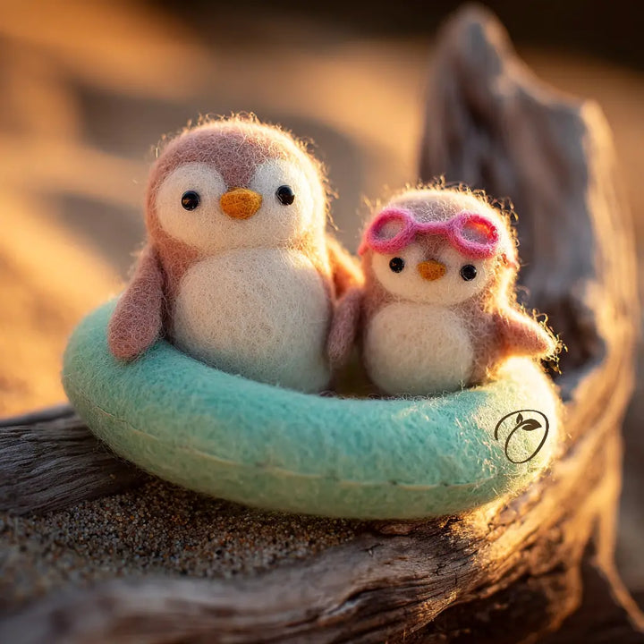 Two small felt penguin toys sit in a mint-green ring on a piece of driftwood outdoors at warm golden hour. One penguin is pink and white, and the other wears pink goggles.