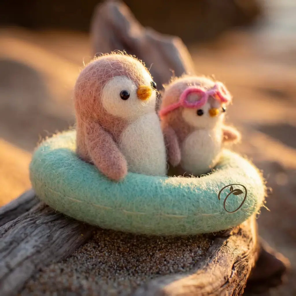 Two small felt penguin figurines sit in a mint-green donut float on a weathered log outdoors. One penguin is pink and white, and the other wears pink goggles.