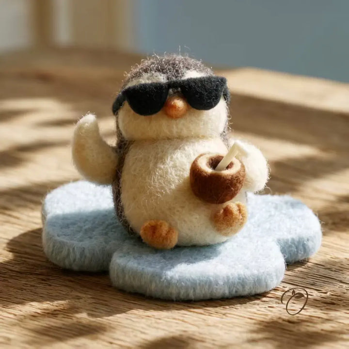 A small felt penguin figurine wearing black sunglasses and holding a brown coconut drink, sitting on a pale blue flower-shaped base on a wooden surface with dappled sunlight.