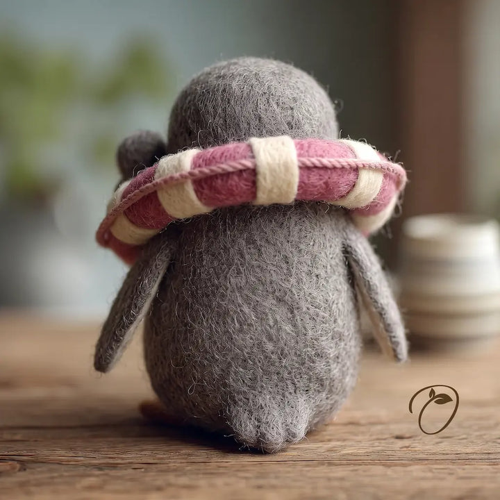 A small gray felt penguin figurine sits on a wooden surface, viewed from behind, wearing a pink and cream striped felt scarf. The background is softly blurred with warm indoor lighting.