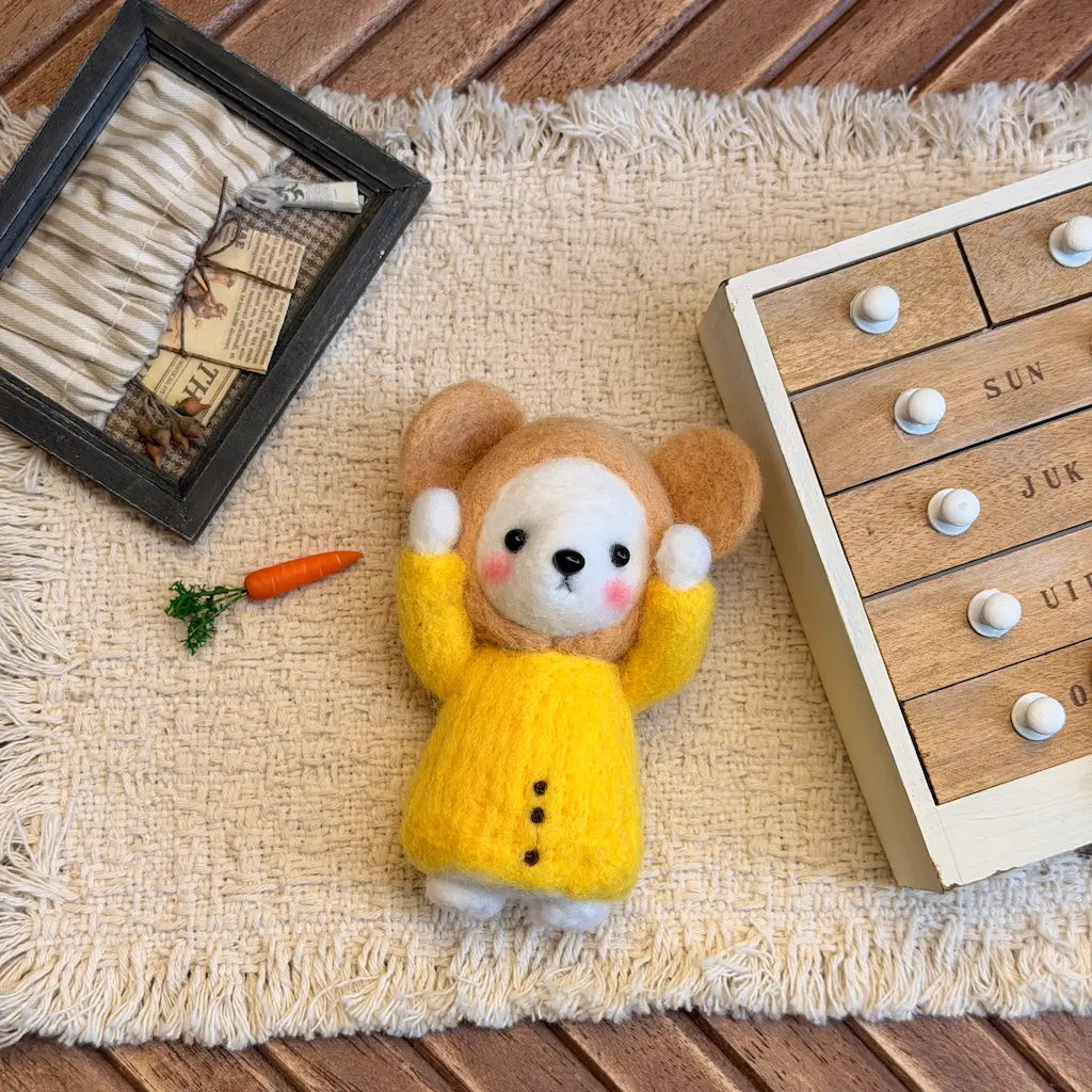 A small felt animal doll with brown ears, a cream face, pink cheeks, and a bright yellow dress, posed on a beige fringed rug with a wooden toy box, a small carrot prop, and a framed dried botanicals item nearby.
