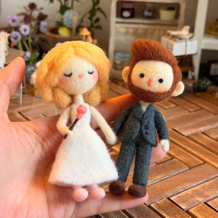A hand holds two small felt wedding figurines: a blonde bride in a white dress with a pink rose, and a brown-haired groom in a gray suit, against a softly blurred indoor background.